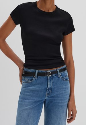 Persoon draagt een aansluitend zwart T-shirt met korte mouwen, in een blauwe spijkerbroek met een zwarte riem, staand met één hand op de heup tegen een effen achtergrond.