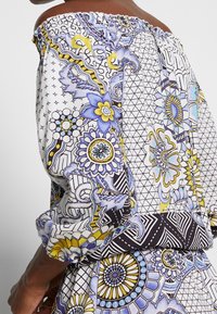 Haut à épaules dénudées avec un motif floral et géométrique vibrant en bleu, jaune et noir, doté d'un col élastique et de poignets élastiqués.