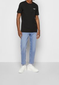 Man som bär en svart kortärmad tröja från Calvin Klein Jeans, ljusblå jeans, vita strumpor och vita sneakers som står mot en vit vägg.