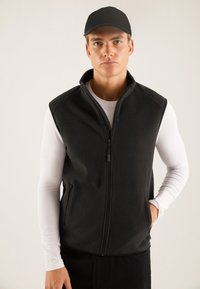 Black fleece vest with a high collar, front zipper, and side pockets, worn over a white long-sleeve shirt. Smooth texture, no patterns.