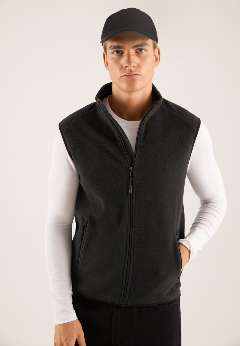 Black fleece vest with a high collar, front zipper, and side pockets, worn over a white long-sleeve shirt. Smooth texture, no patterns.