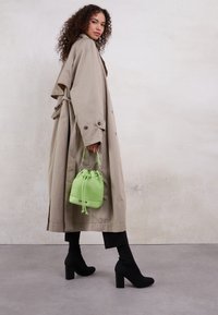 Vaalean beige trench-takki sivunapeilla, yhdistetty mustiin höyhenkorko ankle-saappaisiin. Kirkkaan vihreä ämpärilaukku on pidetty lyhyellä kahvalla.