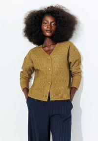 Un cardigan tricoté de couleur moutarde présentant un motif en losanges, un col en V et cinq boutons à l'avant, associé à un pantalon rayé bleu marine.