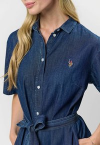 Džínové šaty v tmavomodrej farbe, s krátkymi rukávmi, zapínaním na gombíky a viazaním v páse. Malý vyšívaný detail pri golieri.