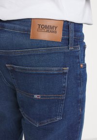 Mörkblå denimjeans med en brun läderlapp märkt "TOMMY JEANS" på baksidan, med orangea sömmar och en liten logotyp på fickan.