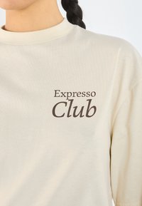 Béžové bavlněné tričko s krátkým rukávem s tmavě hnědým textovým grafickým motivem "Expresso Club" na levém prsu.