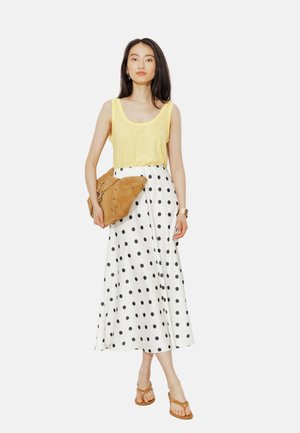 Femme portant un haut jaune sans manches, une jupe midi blanche à pois noirs, des sandales beige et tenant une pochette beige.