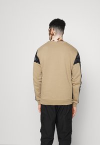 Sudadera beige con acentos en los hombros de patrón negro, puños y borde acanalados; presenta un cuello redondo y un corte relajado. Se llevan pantalones negros por debajo.