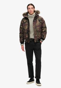 Kamuflage pufferjakke med pelskantet hætte, grå sweater, sorte bukser og sorte sneakers med hvide såler. Har lynlås og lommer.