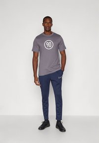 Siva majica s bijelim okruglim dizajnom "90", u kombinaciji s tamnoplavim hlačama s malim Nike logotipom. Model nosi crne tenisice.