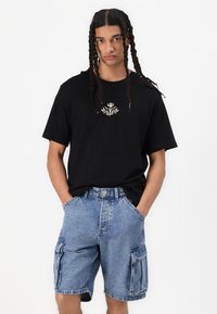 T-shirt in cotone nero con ricamo floreale bianco sul petto, abbinato a pantaloni cargo in denim blu con tasche laterali.