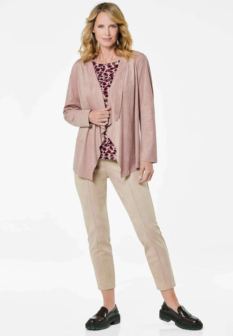 Helles rosa, drapierter Cardigan über einem gemusterten Oberteil, kombiniert mit beigen Hosen und schwarzen Slippern. Geschmeidige Textur mit lockerer Passform.