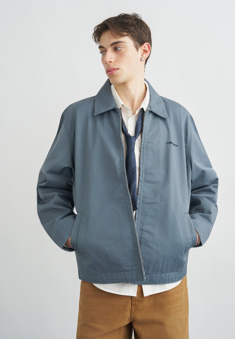 Giovane uomo che indossa una giacca blu con cerniera, camicia bianca, cravatta blu scuro e pantaloni marroni, in piedi con le mani in tasca che guarda a sinistra.