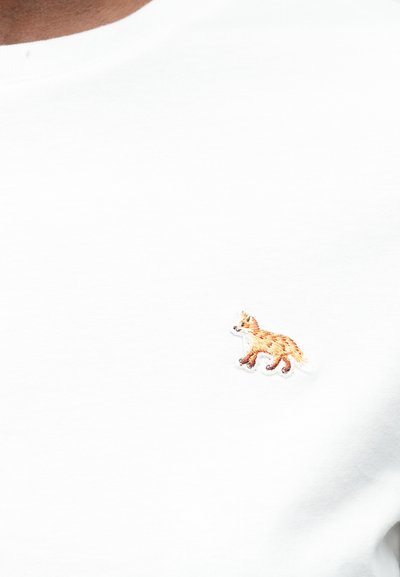 Malá vyšitá oranžová liška na hrudi jednoduchého bílého trička, které má na sobě osoba.