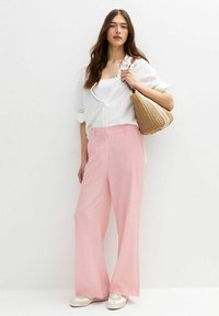 Lichtroze wijde broek van zachte stof, gecombineerd met een witte overhemdblouse en een strooien tas, gedragen met witte sneakers.