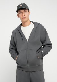 Felpa grigia con zip realizzata in tessuto morbido, dotata di tasche a marsupio, cappuccio con coulisse e un piccolo logo sul petto e sul polsino.