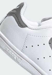 White leather sneaker with a textured finish, grey accents on the heel, perforated details on the sides, and a rubber sole with logo.