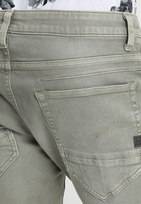 Jeans en denim vert olive clair avec une finition texturée, dotés de poches arrière, de coutures contrastantes et d'une petite étiquette de marque sur la poche droite.