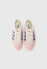 Różowe zamszowe sneakersy z granatowymi paskami, białymi sznurowadłami i beżową gumową podeszwą. Szczegóły obejmują przeszywane akcenty oraz logo marki.