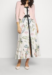 Abito maxi floreale in tessuto bianco, con foglie verdi e dettagli rosa. Abbinato a un cardigan corto rosa chiaro e una cintura nera.