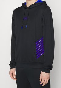 Svart hoodie med blå accenter, som har en logotyp, en framficka och randiga lila detaljer på sidan. Mjuk, texturerad tyg.