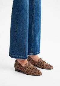 Leopard print loafers made of suede, featuring a brown base with black spots and a gold accent on the strap. Paired with wide-leg blue jeans.
