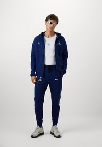 Marineblå træningssæt med Nike-logo, der inkluderer en zip-up hoodie og bukser med elastisk talje. Indeholder broderede detaljer og grå sportssko.