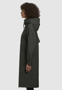 Giacca lunga, di un verde scuro impermeabile, con cappuccio, vita stretta e tasche con zip. Presenta polsini elastici e un logo sulla manica.