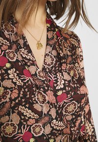 Tissu noir à motifs floraux avec des accents rouges et roses, avec un design en V. Un collier en pendentif doré avec un gland est porté.