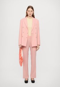 Pink tailored suit with double-breasted blazer and straight-leg trousers; paired with a light yellow top and a pink satin handbag. Black shoes.