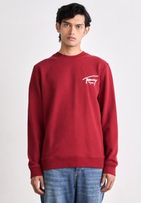 Giovane uomo che indossa un felpa rossa di Tommy Jeans e jeans blu, in piedi su uno sfondo bianco semplice.