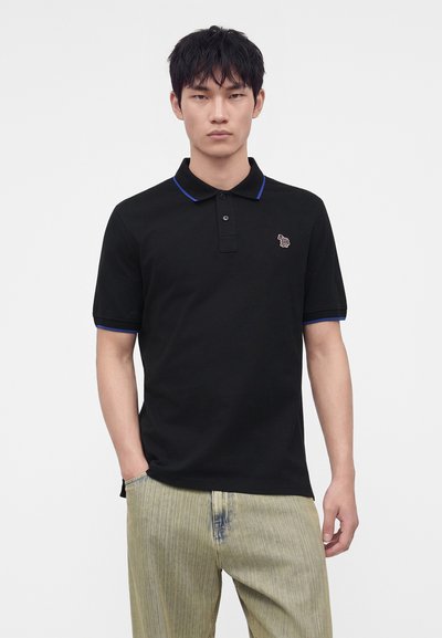 Jeune homme portant un polo noir avec un liseré bleu et un petit logo brodé, debout, une main dans la poche, sur fond blanc.
