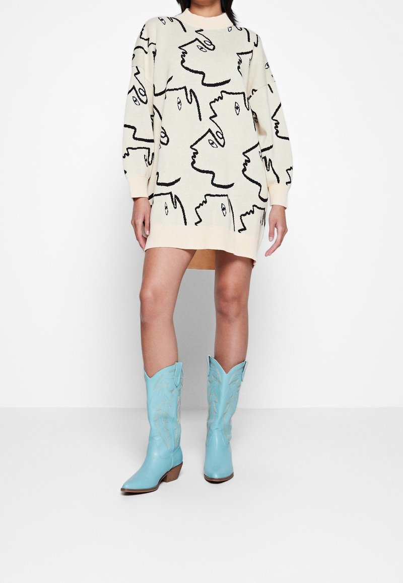 Femme portant une robe-pull crème avec un dessin abstrait de visage en ligne noire et des bottes de cowboy bleu clair, debout devant un fond blanc uni.