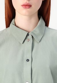 Camicia in velluto a coste verde con colletto, quattro bottoni frontali e texture a coste visibile. Vista ravvicinata della parte superiore del capo.
