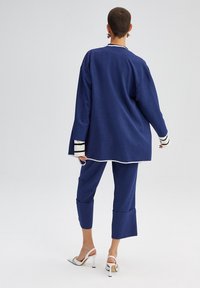 Volno střižená námořnicky modrá bunda s dlouhými rukávy a bílým lemováním, doplněná odpovídajícími zkrácenými kalhotami. Obuv má bílé podpatky.