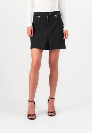 Vrouw draagt een zwarte minirok met een ritssluiting aan de voorkant en gouden ritssluitingszakken, gecombineerd met zwarte sandalen met bandjes en een wit top met lange mouwen.