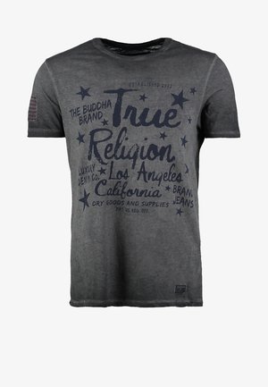 Mann trägt ein dunkelgraues True Religion Grafik-T-Shirt, dunkelblaue Jeans, schwarze Schuhe und einen schwarzen Schultertaschengurt quer über der Brust.