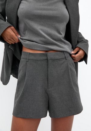 Person iført grå skræddersyede shorts og matchende blazer, løfter den grå top for at afsløre taljen, mod en ensfarvet lys baggrund.