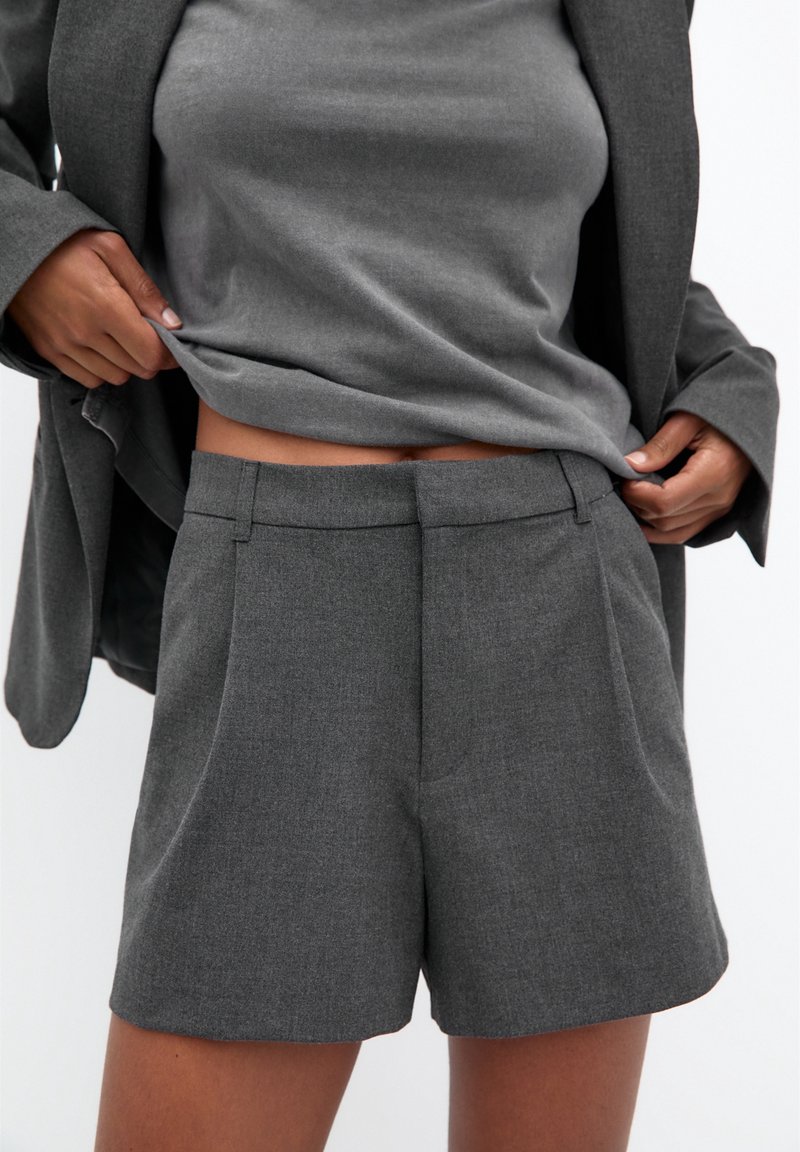 Personne portant un short gris sur mesure et un blazer assorti, soulevant un haut gris pour révéler la taille, sur un fond clair uni.