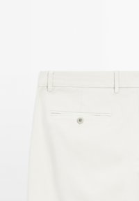 Light beige trousers with a smooth texture, featuring a back pocket with a button closure and belt loops. Minimalist design.