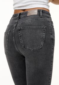 Person iført høj talje mørkegrå jeans med baglommer og et synligt lædermærke på taljeribben, kombineret med en kort hvid top.