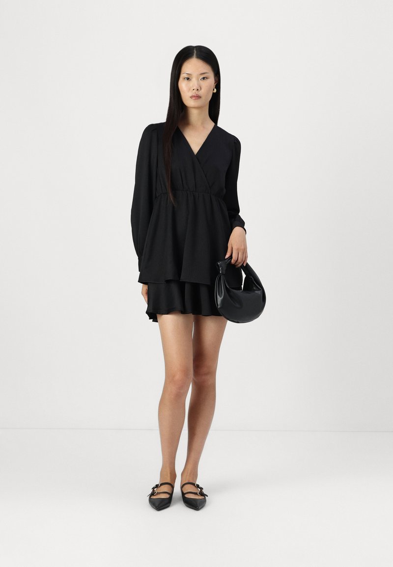 Robe noire enveloppante à manches longues, design superposé, texture douce ; assortie à un petit sac à main noir et des chaussures noires à bouts pointus avec des lanières.