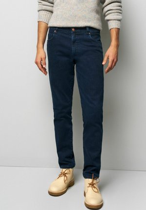 Dunkelblaue Jeans mit schlankem Schnitt und fünf Taschen, ausgestattet mit einem kupferfarbenen Knopf und kontrastierenden Nähten. Getragen mit hellen Stiefeln.