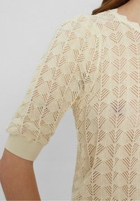 Světle krémový pletený svetr s vzorem listů, krátké nabírané rukávy a vroubkované manžety. Lehký a strukturovaný materiál s otevřeným tkaním.