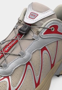 Μπεζ και κόκκινο αθλητικό παπούτσι με ύφασμα από δίχτυ, συνθετικά επικάλυψης, κορδόνια γρήγορου σφιξίματος και μαύρο πλαστικό κλείδωμα κορδονιών στην κορυφή.