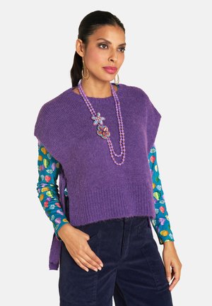 Lila gestrickte Poncho mit seitlichen Schlitzen, über einem bunten gemusterten Hemd getragen, ergänzt durch eine mit Perlen besetzte Blumenhalskette und dunkle Hosen.