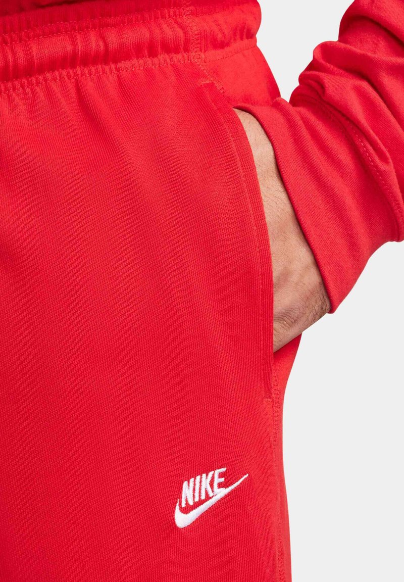 Czerwone sportowe dresy z elastycznym paskiem w talii, bocznymi kieszeniami i białym logo Nike. Materiał wydaje się gładki i lekko elastyczny.