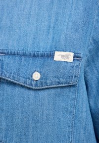 Jasnoniebieska koszula dżinsowa z guzikową kieszenią na piersi, białym zapięciem na guziki oraz małą wplecioną metką z szarym wzorem.