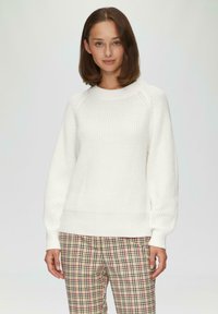 Witte gebreide trui met een ribbelstructuur, ronde hals en losse mouwen, gecombineerd met een geruite broek in beige, rood en groene tinten.