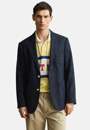 Homme portant un blazer bleu foncé, un polo jaune avec des rayures bleues et rouges, et un pantalon beige sur un fond clair uni.